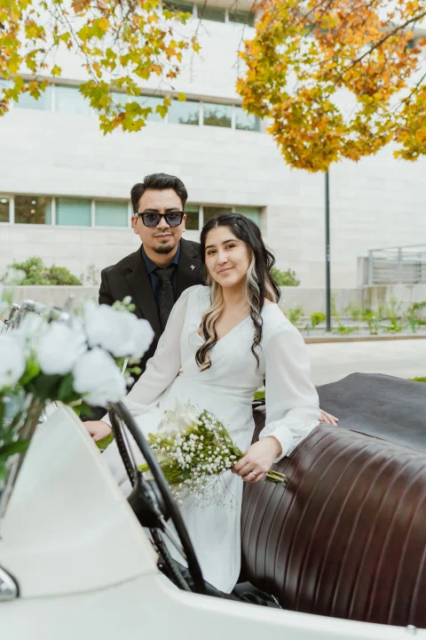 Mujer con vestido blanco está sentada en un automóvil convertible blanco con un ramo, mientras un hombre permanece junto al vehículo.