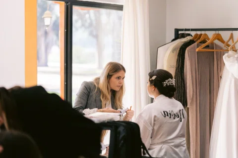 Mujer sentada junto a una ventana recibe maquillaje mientras lleva el cabello recogido.