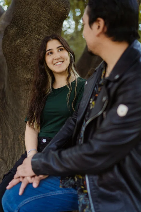 Mujer sonriendo al hombre mientras ambos están sentados junto al tronco de un árbol.