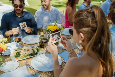 Mujer sostiene una fotografía instantánea junto a una mesa preparada donde varias personas están sentadas.