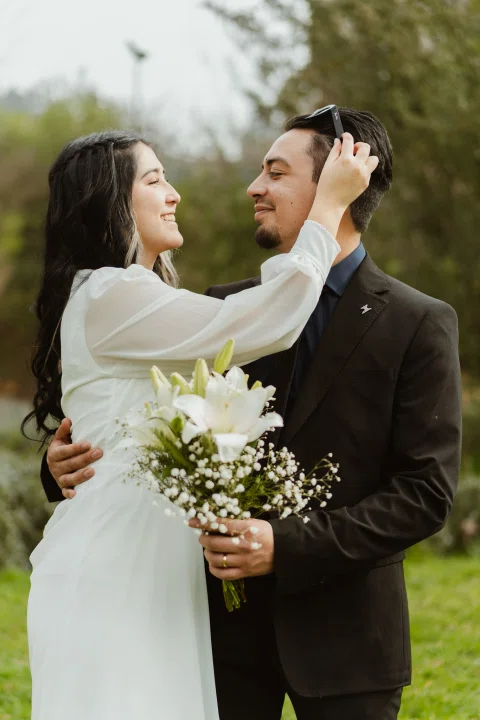 Mujer con vestido blanco toca el cabello de un hombre que sostiene un ramo.