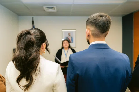 Mujer con traje blanco y hombre con traje azul permanecen de espaldas frente a una mujer situada detrás de un escritorio.