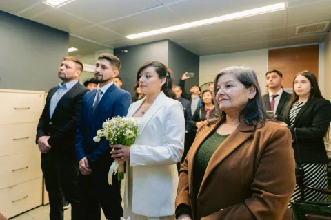 Mujer con traje blanco sostiene un ramo entre varias personas dentro de una sala.