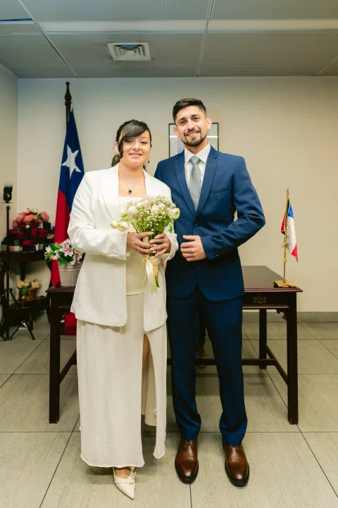 Mujer con traje blanco sostiene un ramo junto a un hombre con traje azul dentro de una sala.