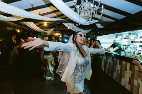 Mujer con traje claro y lentes oscuros baila con los brazos extendidos frente a una barra.