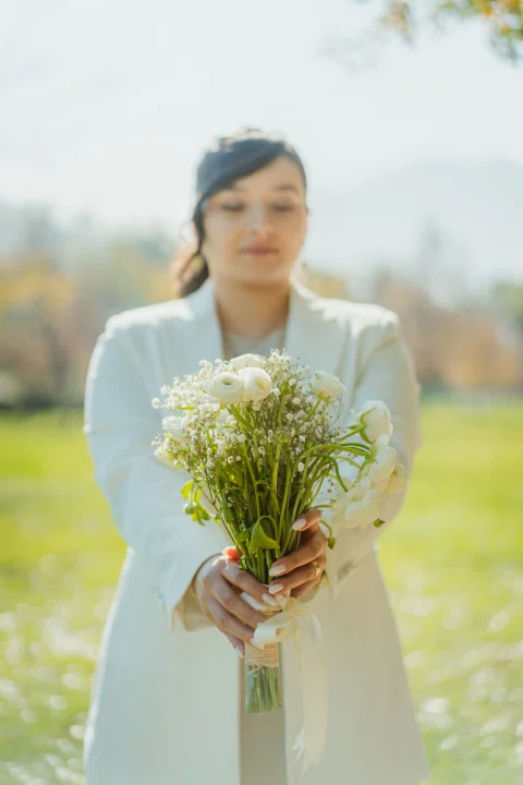 Mujer con traje claro sostiene un ramo de flores frente a la cámara en un parque.
