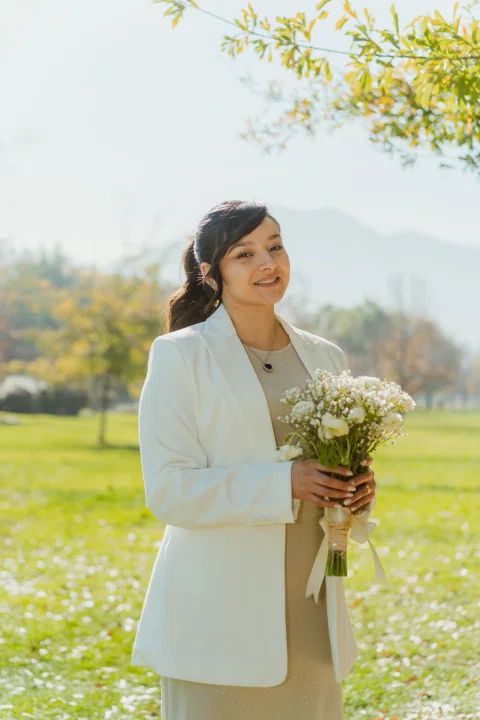 Mujer con traje claro sostiene un ramo de flores en un parque, con vegetación desenfocada al fondo.