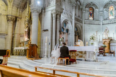 Mujer con velo y hombre con traje oscuro permanecen frente a una persona con vestimenta religiosa, vistos desde un costado de la iglesia.