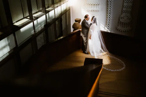 Mujer con velo y hombre con traje oscuro permanecen juntos en una escalera interior iluminada a contraluz.