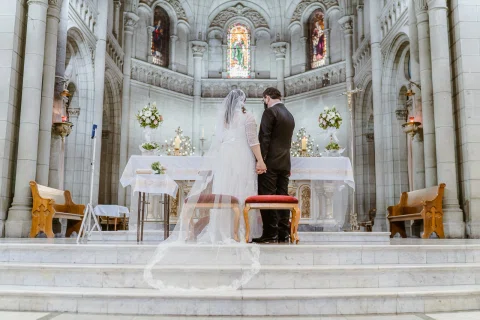 Mujer con velo y hombre con traje oscuro permanecen tomados de la mano frente al altar de una iglesia.