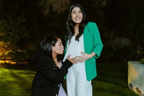 Mujer vestida de negro besa el abdomen de una mujer con blazer verde que sonríe en un jardín de noche.