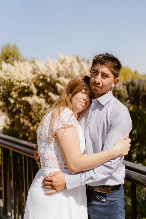 Mujer con vestido blanco abraza a un hombre con camisa celeste junto a una baranda en un parque.