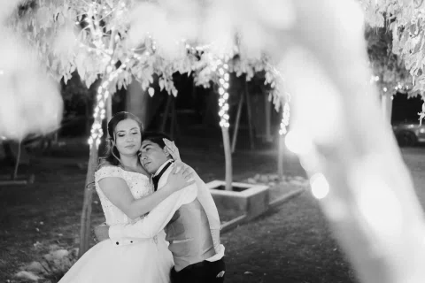Mujer con vestido blanco abraza a un hombre con chaleco claro bajo luces nocturnas, en una fotografía en blanco y negro.
