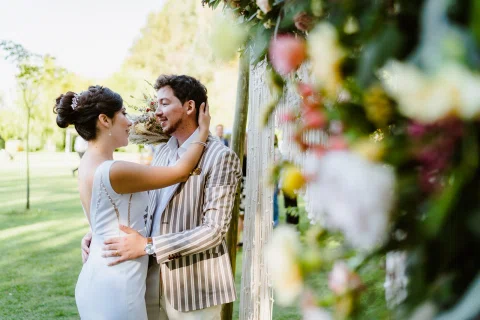 Mujer con vestido blanco abraza a un hombre con chaqueta a rayas detrás de flores desenfocadas en primer plano.