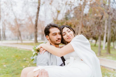 Mujer con vestido blanco abraza a un hombre con traje claro mientras él la sostiene en brazos en un parque.