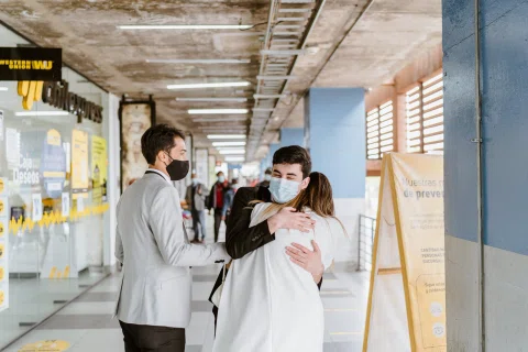 Mujer con vestido blanco abraza a un hombre con traje oscuro y mascarilla, mientras un hombre con traje claro permanece a su lado en un corredor.