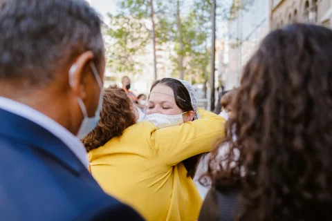 Mujer con vestido blanco abraza a una mujer vestida de amarillo a la salida de una iglesia.