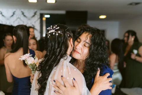 Mujer con vestido blanco abraza a una mujer con vestido azul entre varias personas.