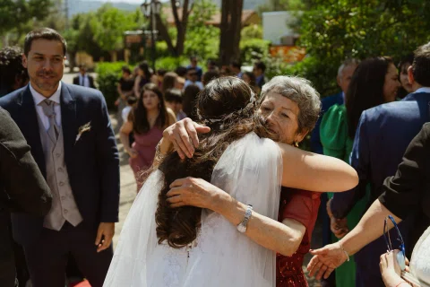 Mujer con vestido blanco abraza a una mujer de cabello canoso en el exterior de la capilla.