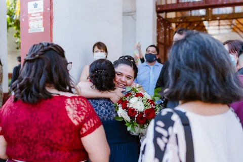 Mujer con vestido blanco abraza a otra mujer en el exterior mientras varias personas las rodean.