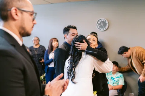 Mujer con vestido blanco abraza a otra mujer mientras un hombre calvo aparece en primer plano.