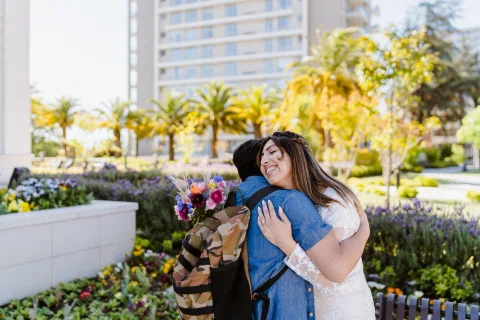 Mujer con vestido blanco abraza a otra mujer en el jardín mientras sostiene un ramo de flores.