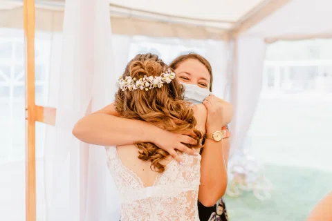Mujer con vestido blanco abraza estrechamente a otra mujer con mascarilla.