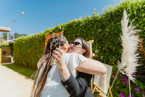Mujer con vestido blanco abraza a una mujer con ropa negra y lentes de sol al aire libre.