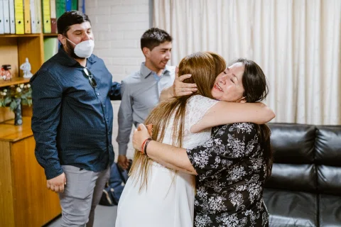 Mujer con vestido blanco abraza a una mujer con vestido estampado dentro de una sala.