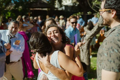 Mujer con vestido blanco abraza a una mujer con vestido rosado entre varias personas.