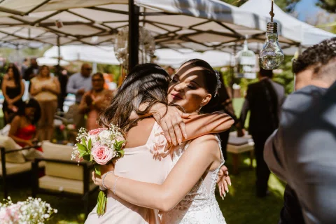 Mujer con vestido blanco abraza a una mujer con vestido rosado en el jardín.