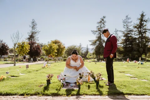 Mujer con vestido blanco se agacha junto a flores y lápidas mientras un hombre con chaqueta burdeos permanece de pie.