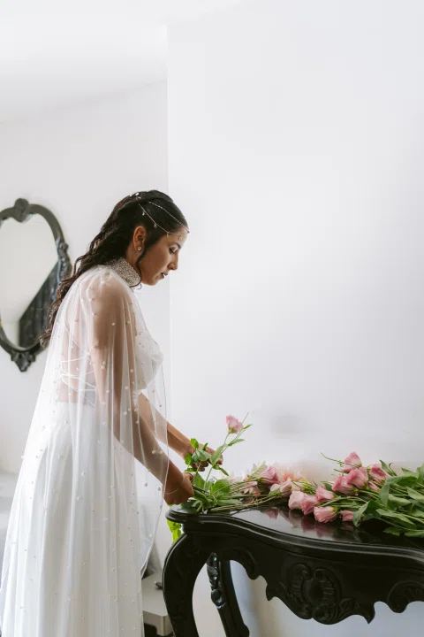 Mujer con vestido blanco acomoda flores sobre una mesa junto a una ventana.