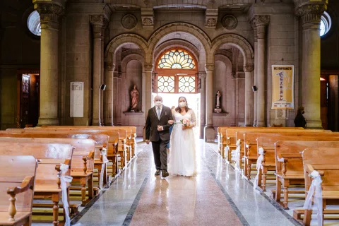 Mujer con vestido blanco avanza por el pasillo de una iglesia junto a un hombre con traje oscuro.