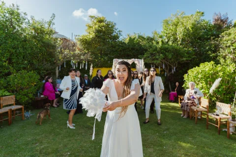 Mujer con vestido blanco avanza sonriendo con un ramo blanco en las manos mientras un grupo permanece detrás.