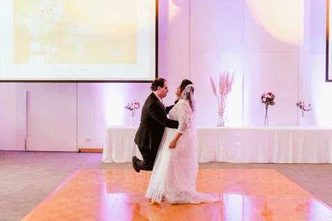 Mujer con vestido blanco baila con un hombre con traje oscuro en un salón iluminado con luces de colores.