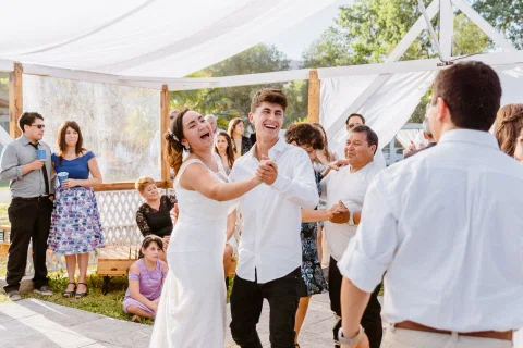 Mujer con vestido blanco baila sonriente con un joven de camisa blanca mientras varias personas los rodean.