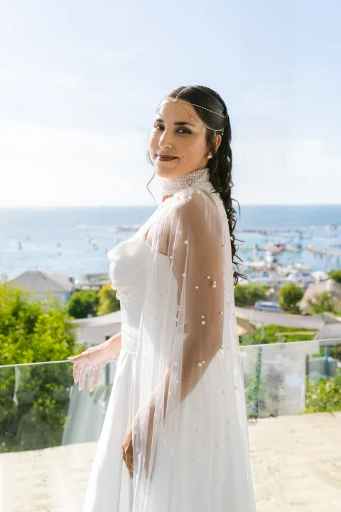 Mujer con vestido blanco posa de perfil junto a una baranda con vista al mar.