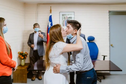 Mujer con vestido blanco besa la mejilla de un hombre con camisa celeste dentro de una sala.