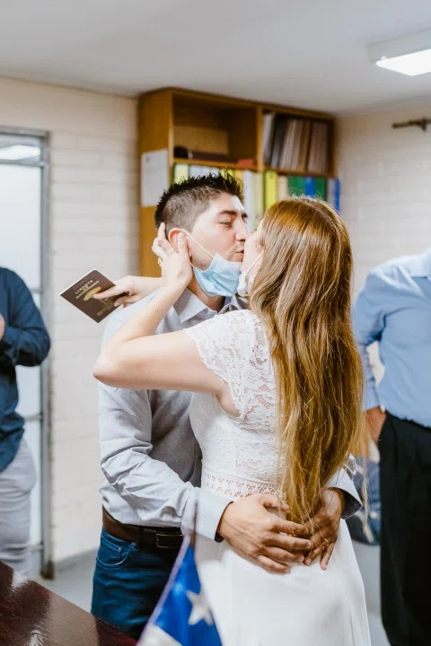 Mujer con vestido blanco abraza y besa la mejilla de un hombre con camisa celeste dentro de la sala.