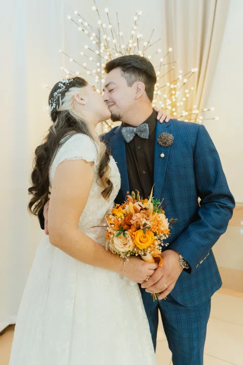 Mujer con vestido blanco besa la mejilla de un hombre con traje azul mientras ambos sostienen un ramo de flores naranjas.
