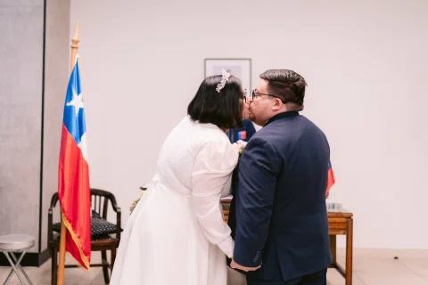Mujer con vestido blanco besa la mejilla de un hombre con traje oscuro junto a una bandera chilena.