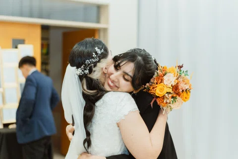 Mujer con vestido blanco abraza y besa la mejilla de otra mujer mientras sostiene un ramo de flores naranjas.