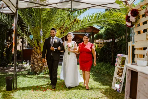 Mujer con vestido blanco camina por el jardín entre un hombre con traje oscuro y una mujer con vestido rojo.