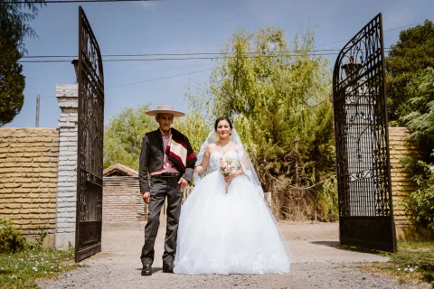 Mujer con vestido blanco camina por un portón junto a un hombre con traje oscuro y sombrero de ala ancha.