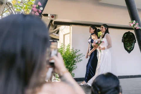 Mujer con vestido blanco camina junto a otra mujer mientras varias personas observan.