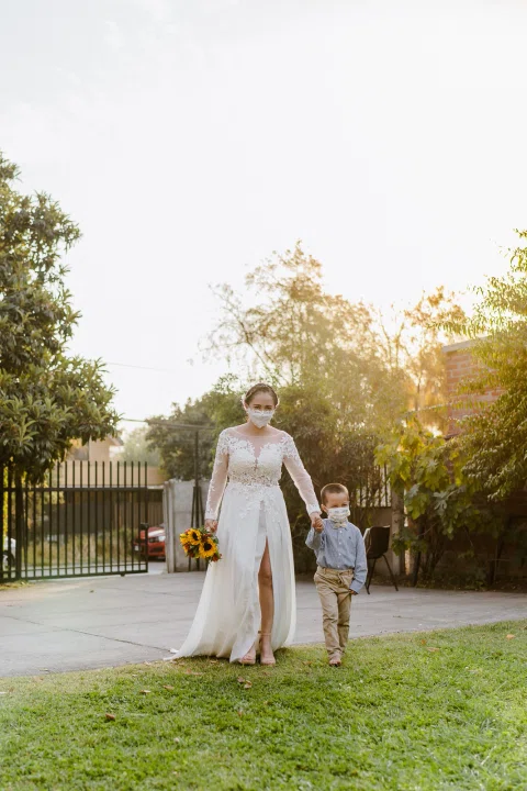 Mujer con vestido blanco camina por el césped junto a un niño; ambos usan mascarilla.