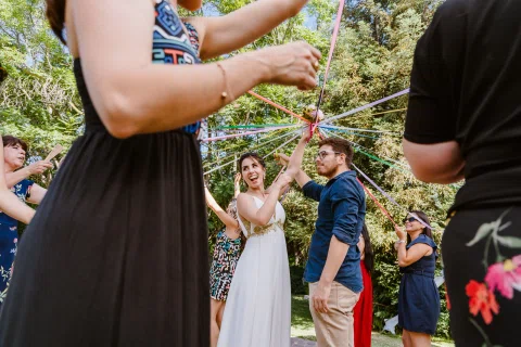 Mujer con vestido blanco permanece en el centro mientras varias personas sostienen cintas de colores que convergen hacia ella durante la celebración.