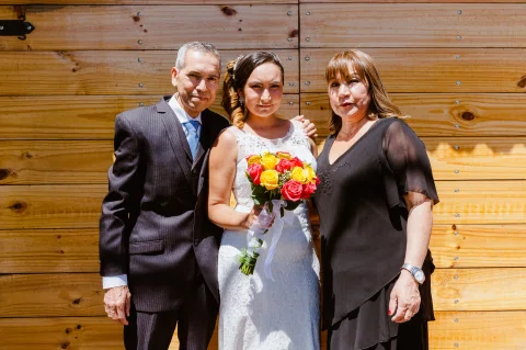 Mujer con vestido blanco posando entre dos adultos frente a un muro de madera.