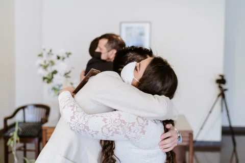 Mujer con vestido blanco y mascarilla abraza a una persona con chaqueta blanca dentro de una sala, mientras otro abrazo ocurre al fondo.